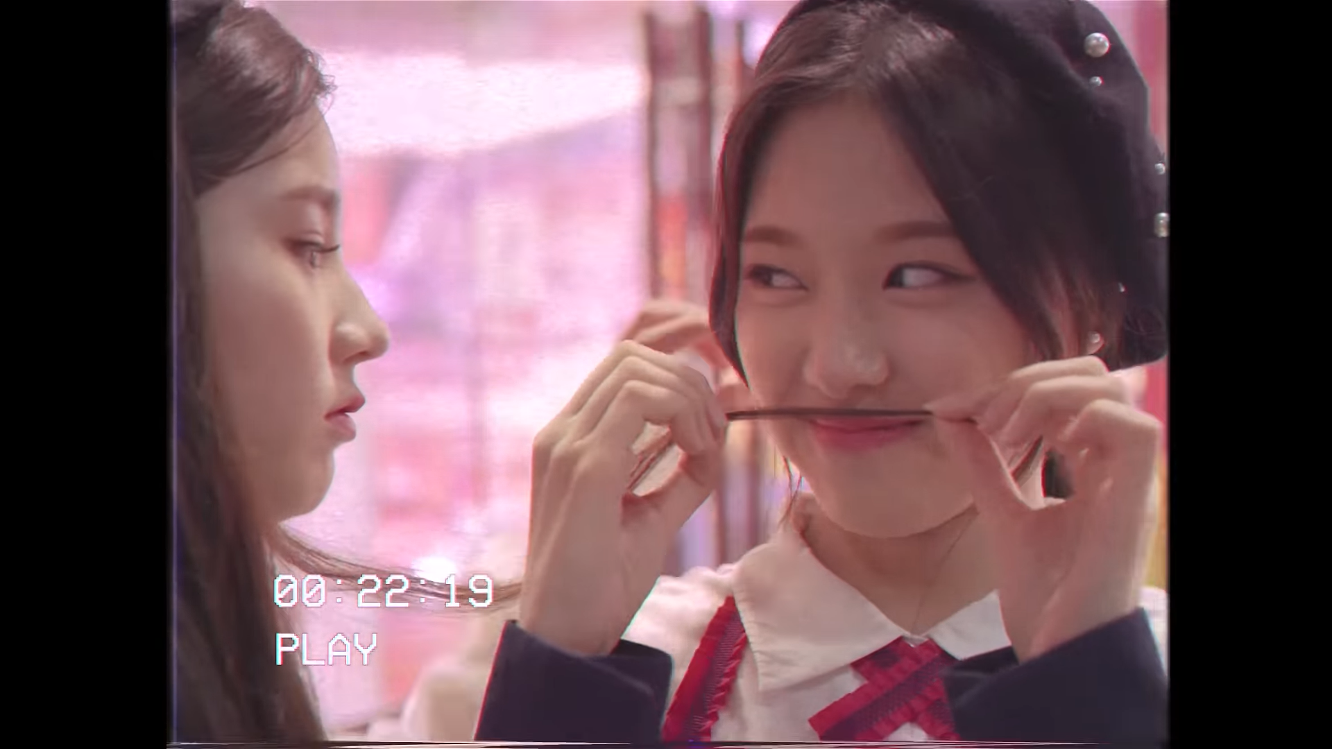 Hyunjin pretending a strand of Heejin's hair is a mustache. There is a filter over the image making it look an old VCR recording. There is white text in the bottom left corner reading '00:22:19' and 'Play.'